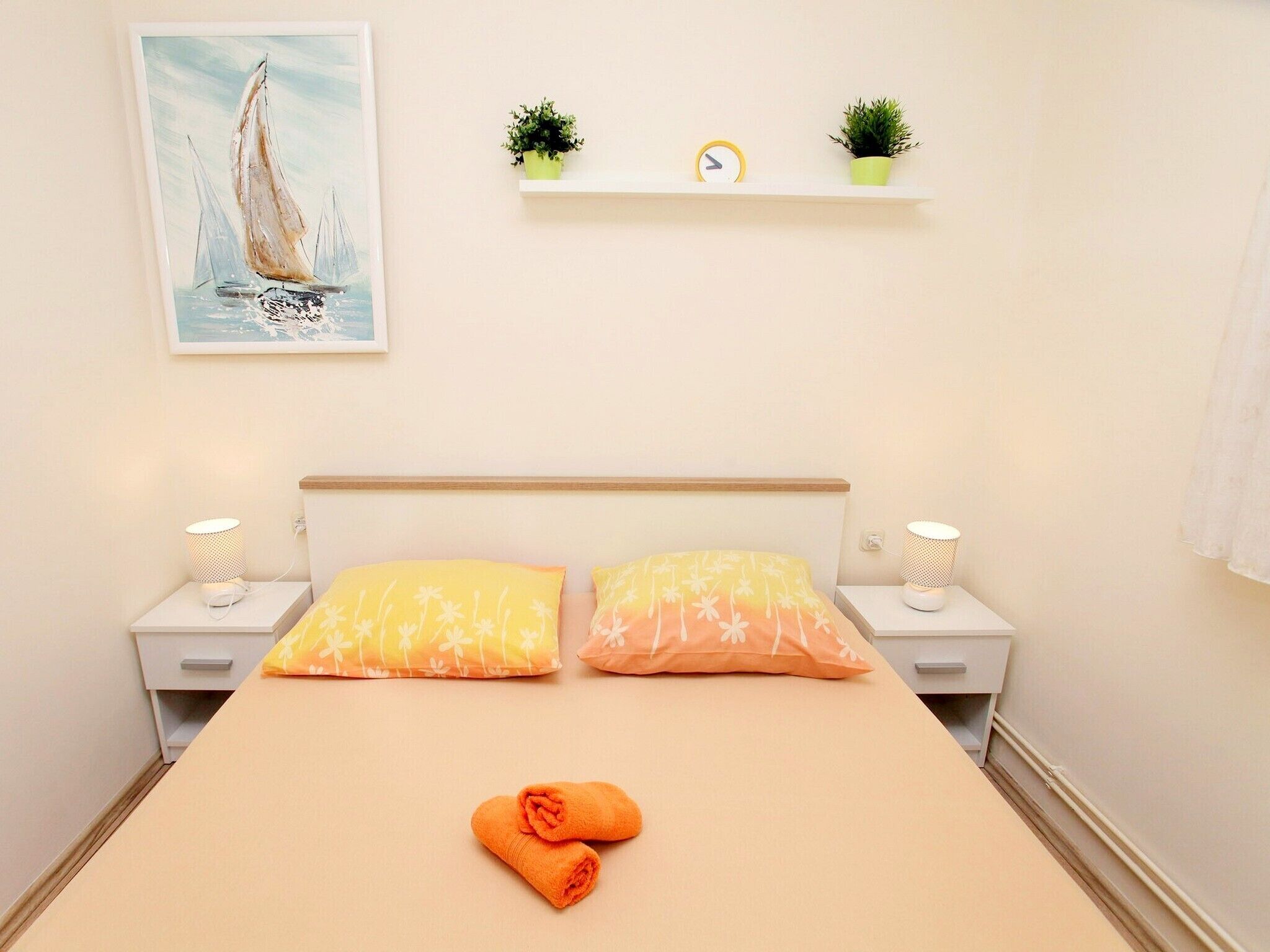 Un dormitorio con una cama, dos mesitas de noche, un estante en la pared con plantas, un reloj y un cuadro de un velero.