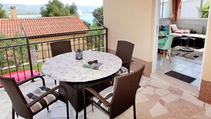 An outdoor patio with a round table and wicker chairs, overlooking a residential area with a tiled roof and a view of the mountains. - Apartments Jozo (Trogir)
