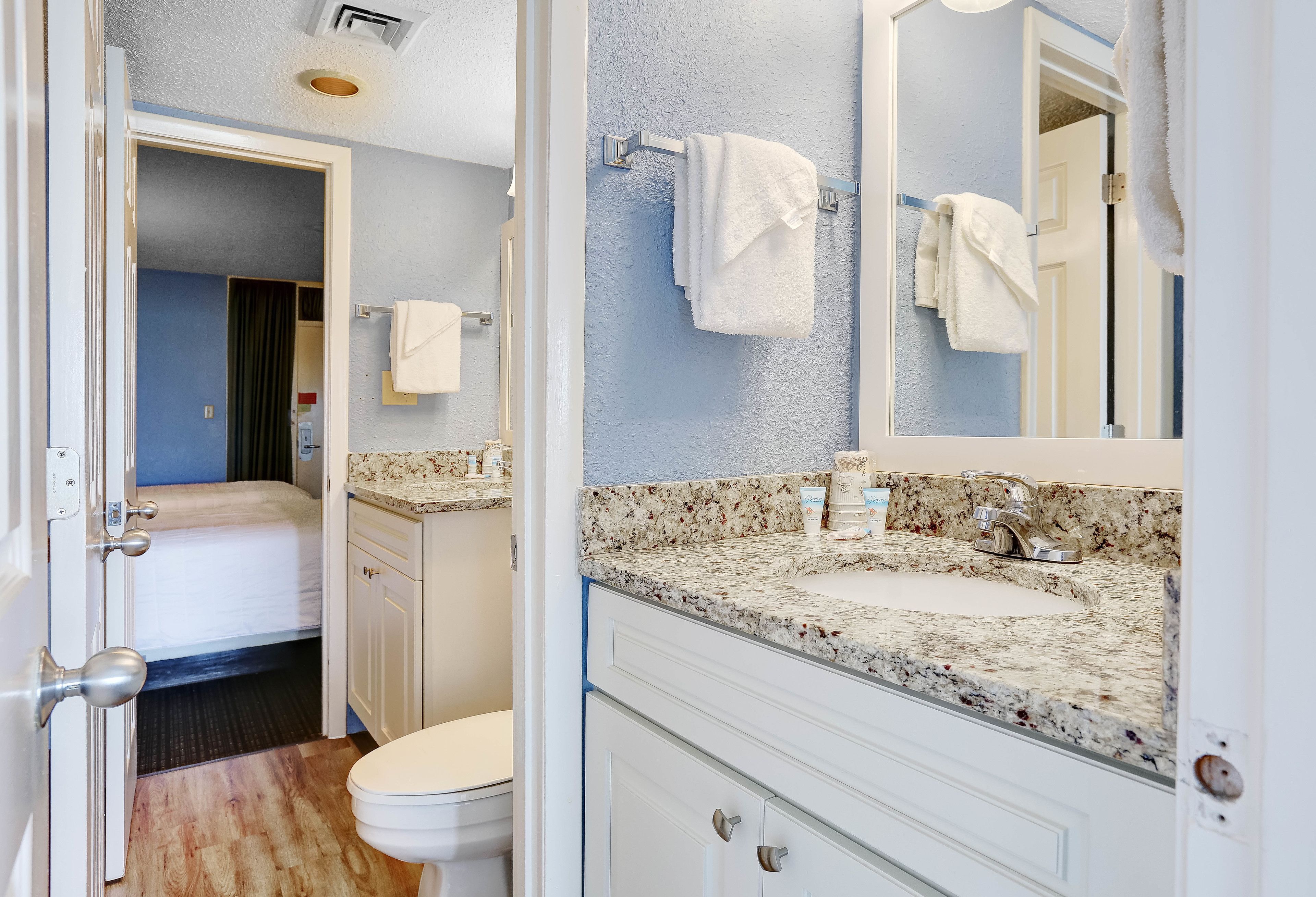 Une salle de bain avec un lavabo en marbre, des meubles blancs et un miroir. Une porte donne accès à une chambre à coucher qui comprend un lit et une salle de bain.