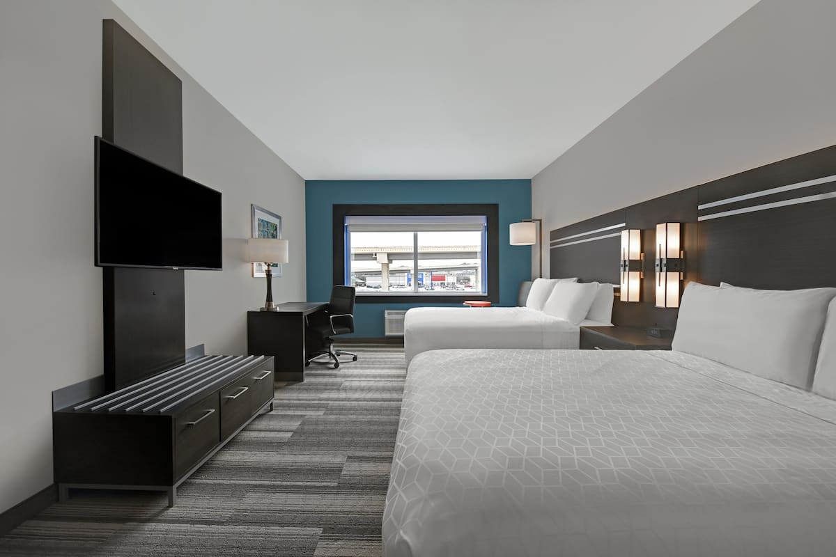 A modern hotel room with a large bed, a desk, a flat-screen TV, and a view of the city through a window.