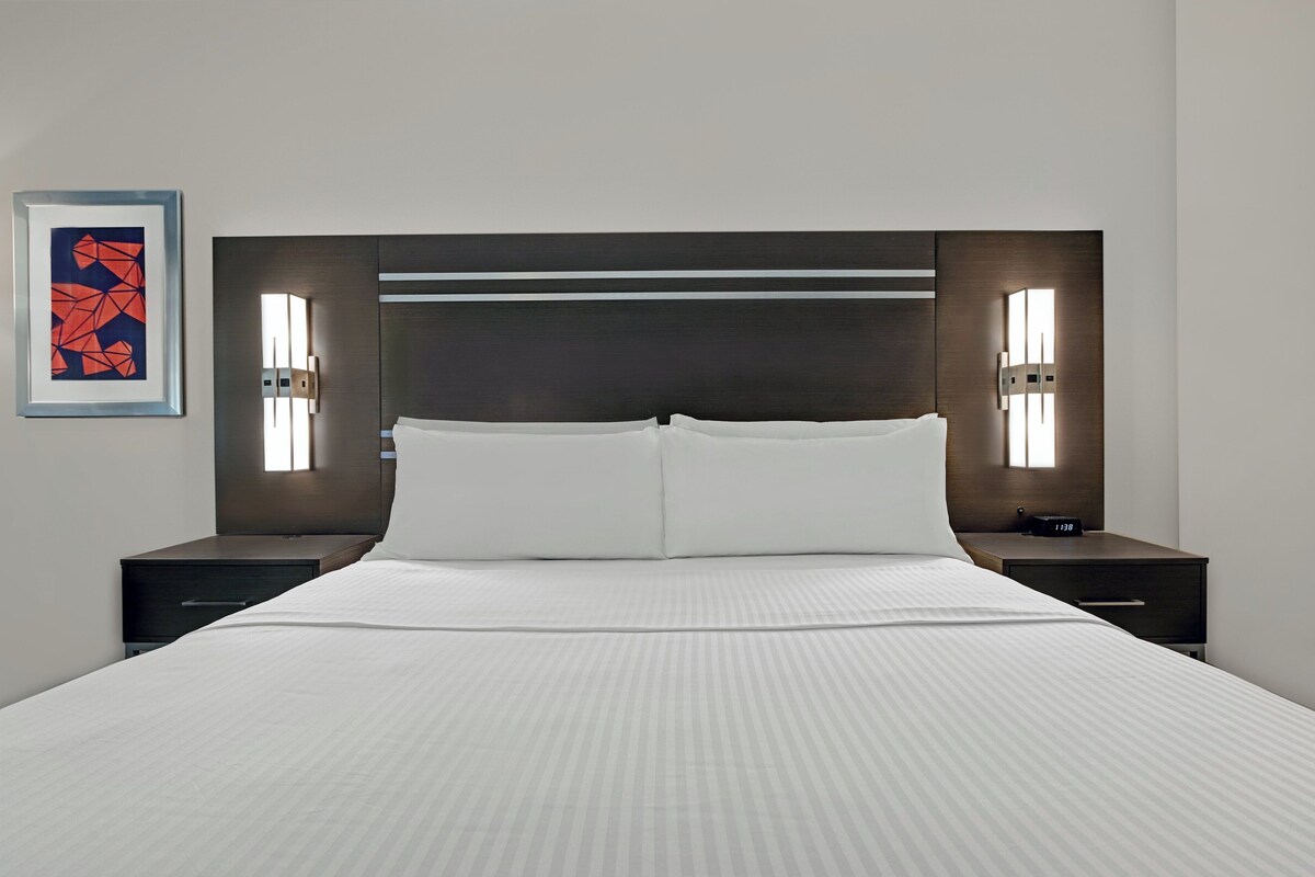 A modern hotel room with a large bed, two bedside tables, a nightstand, and a wall-mounted light fixture.