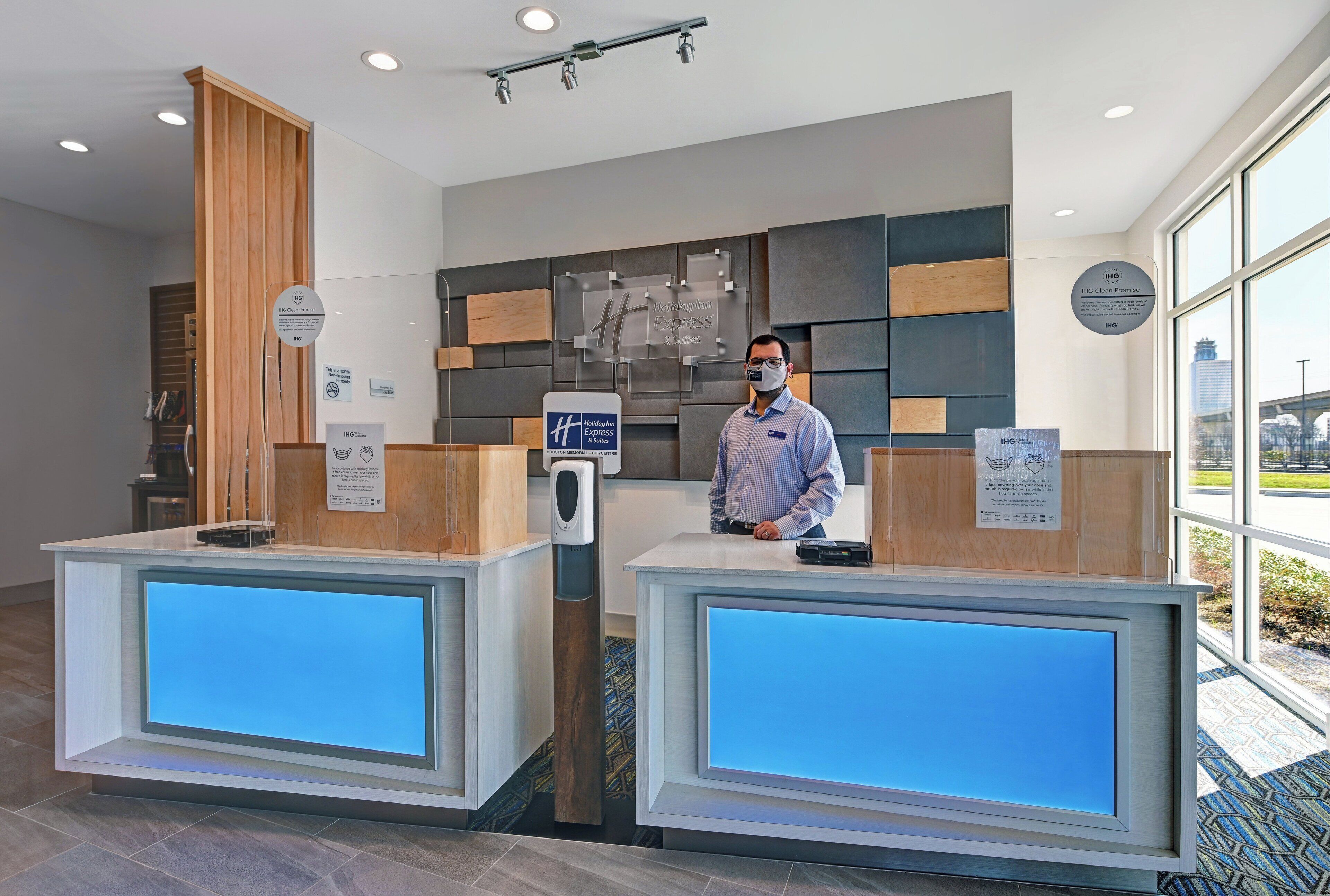A hotel reception desk with a digital display, a hand sanitizer dispenser, and a sign indicating the hotel's commitment to cleanliness.