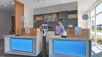 A hotel reception desk with a digital display, a hand sanitizer dispenser, and a sign indicating the hotel's commitment to cleanliness.