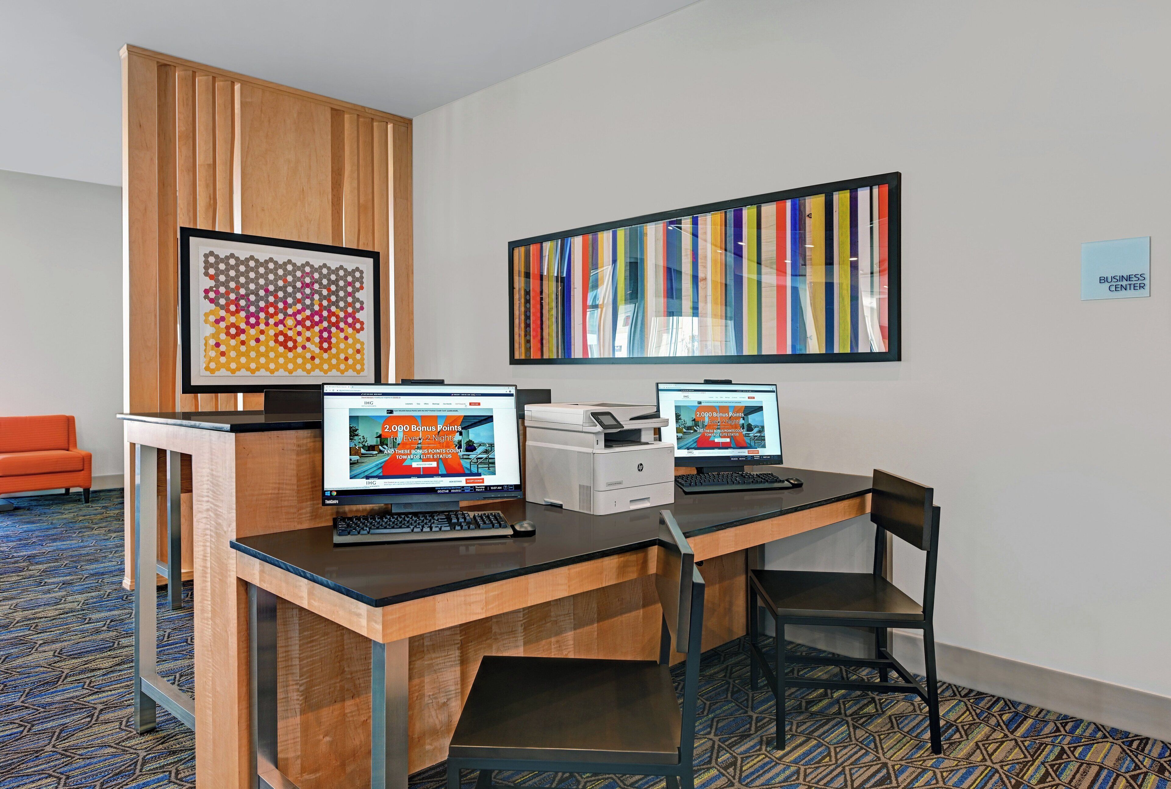 A hotel business center with two computer workstations, a printer, and a large abstract painting on the wall.