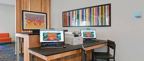 A hotel business center with two computer workstations, a printer, and a large abstract painting on the wall.