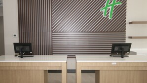 A reception desk with two computer monitors, a wooden desk, and a patterned wall with a green symbol. - Holiday Inn & Suites Fayetteville W-Fort Bragg Area by IHG (Fayetteville)