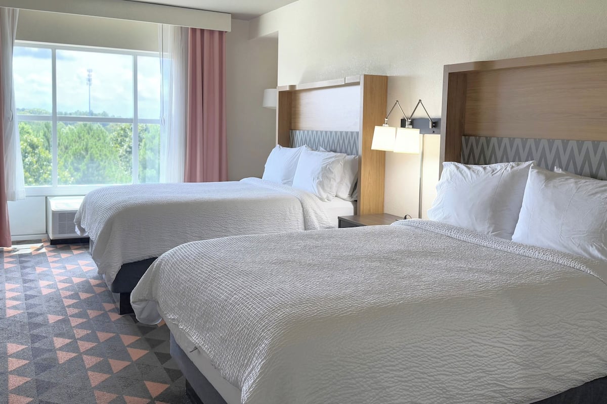 A hotel room with two beds, a large window with a view, a bedside table with a lamp, and a patterned floor.