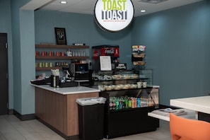 A breakfast counter with a Coca-Cola machine, a glass display case with snacks, and a sign that reads "Toast to Toast Breakfast, Bites & Bar". - Holiday Inn & Suites Fayetteville W-Fort Bragg Area by IHG (Fayetteville)