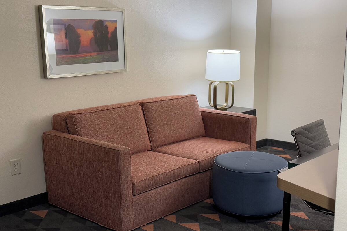 A room with a brown sofa, a round ottoman, a lamp, and a framed painting on the wall.