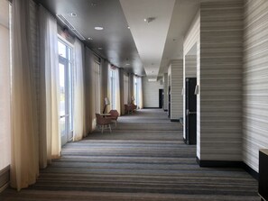 A hotel corridor with a carpeted floor, large windows, and a seating area with chairs and tables.