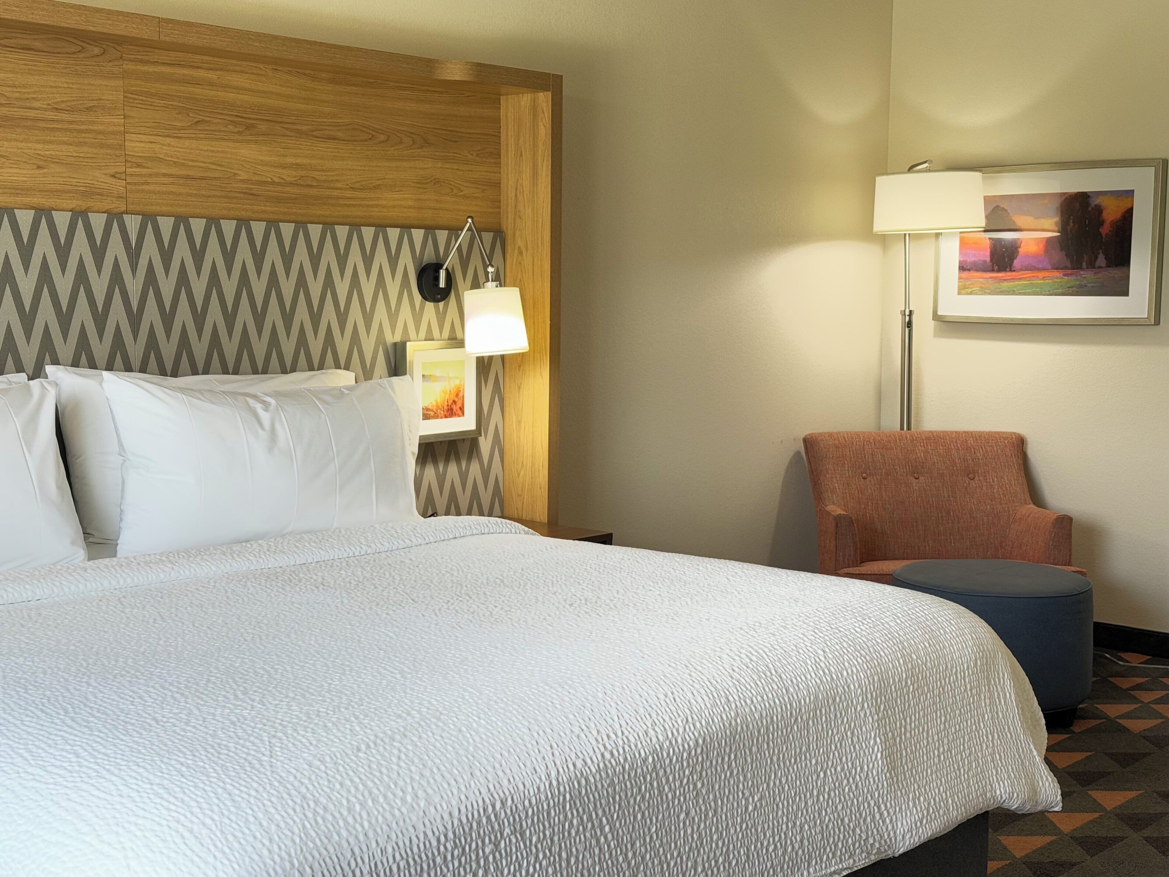 Una habitación de hotel con una cama grande, cabecera de madera, lámpara de pared, un cuadro enmarco, una silla y un pequeño otoman.