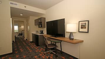 A hotel room with a desk, chair, television, lamp, and kitchenette.