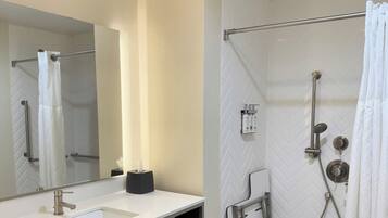A bathroom with a sink, mirror, towel rack, shower, and a towel dispenser.