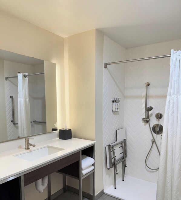A bathroom with a sink, mirror, towel rack, shower, and a towel dispenser.