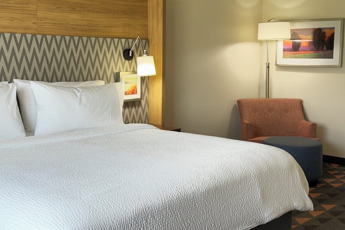 A hotel room with a large bed, a wooden headboard, a wall-mounted lamp, a framed picture, a chair, and a small ottoman.