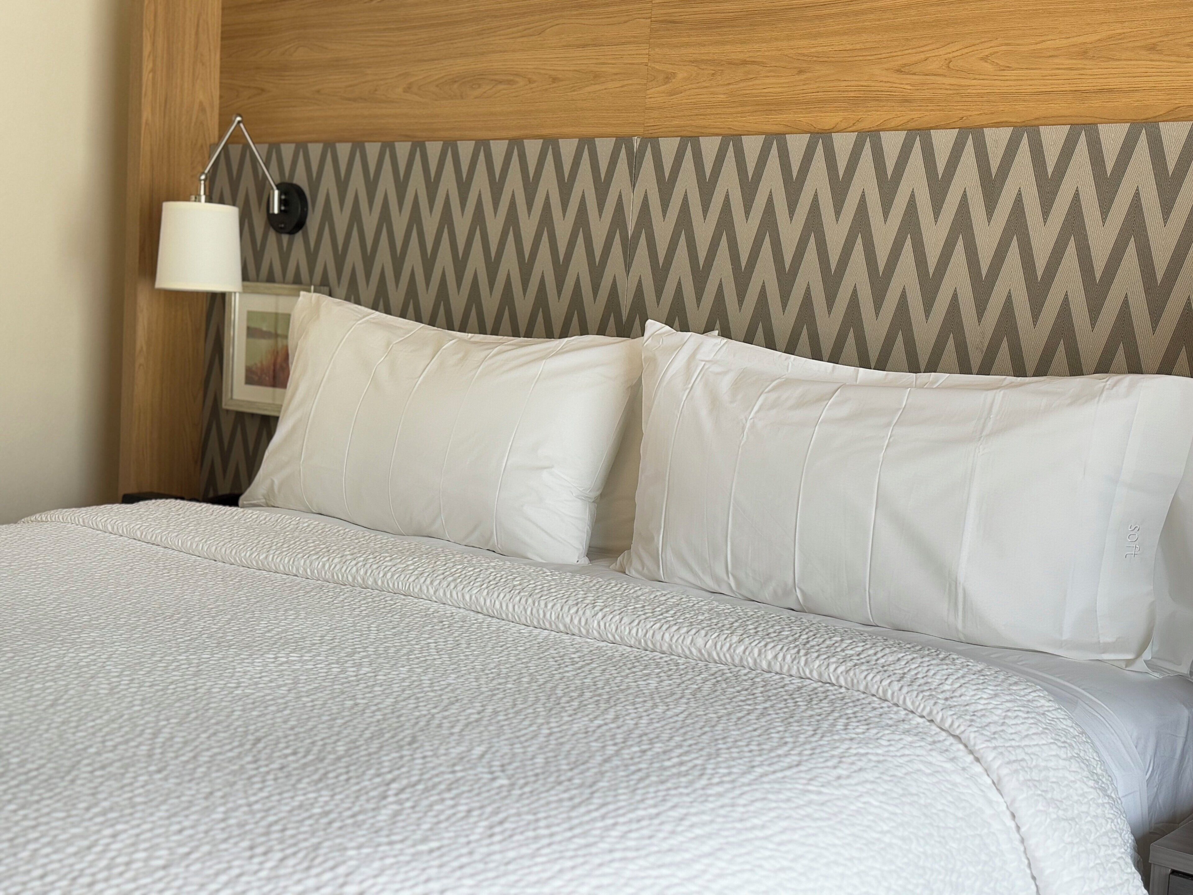 A neatly made bed with white linens and pillows, a wooden headboard, and a wall with a chevron pattern.