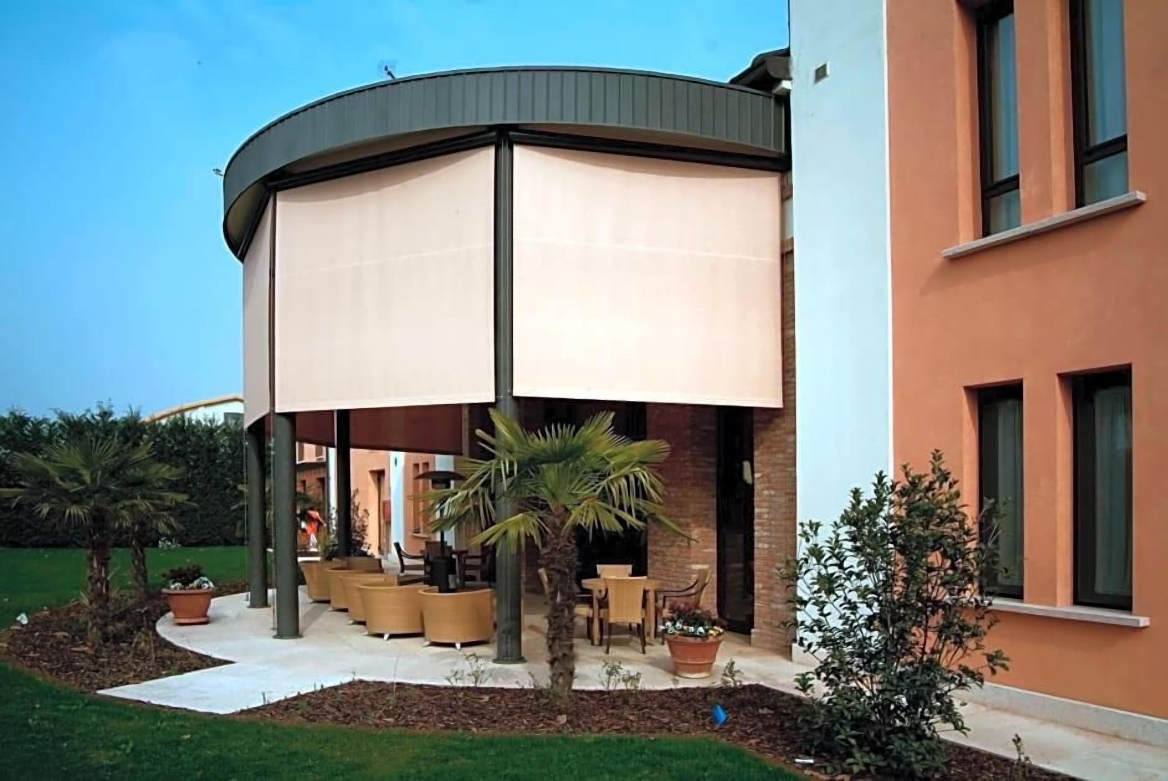 A covered outdoor seating area with tables and chairs, surrounded by plants and a building facade.