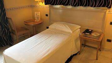 Ein Hotelzimmer mit einem Einzelbett, einem Nachttisch mit Lampe, einem Sessel und einem kleinen Tisch mit Telefon und Papieren.