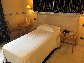 A hotel room with a single bed, a bedside table with a lamp, a chair, and a small table with a phone and papers.