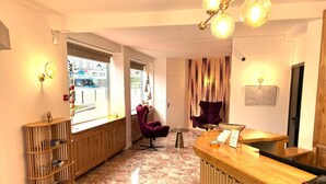 Hall dell'hotel con reception, due poltrone viola, una libreria in legno e un pavimento in pietra.