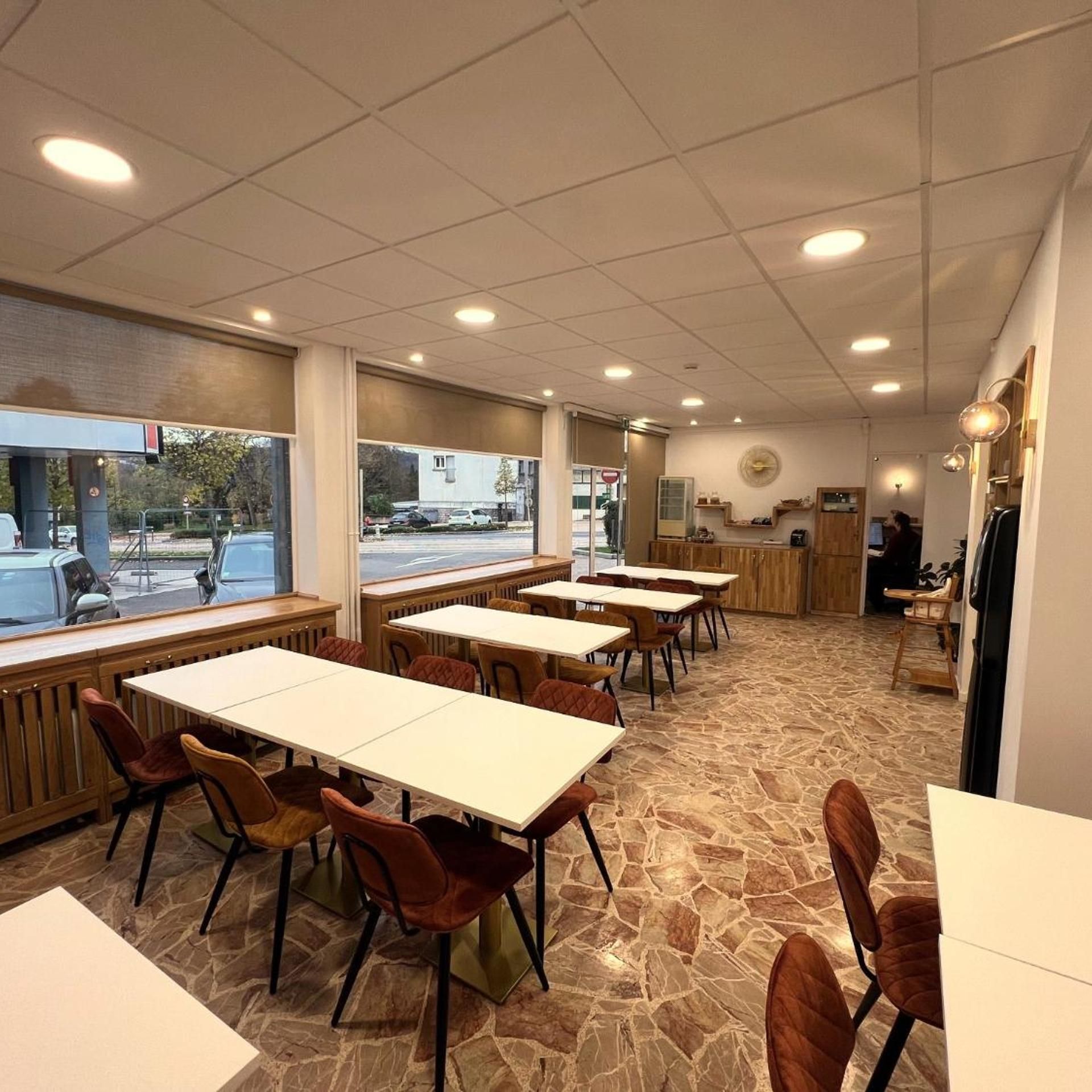 An indoor dining area with tables and chairs, large windows, and a stone floor.
