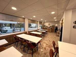 A restaurant with multiple tables and chairs, a stone floor, and a view of the street through large windows. - Contact Hôtel Foch (Besancon)