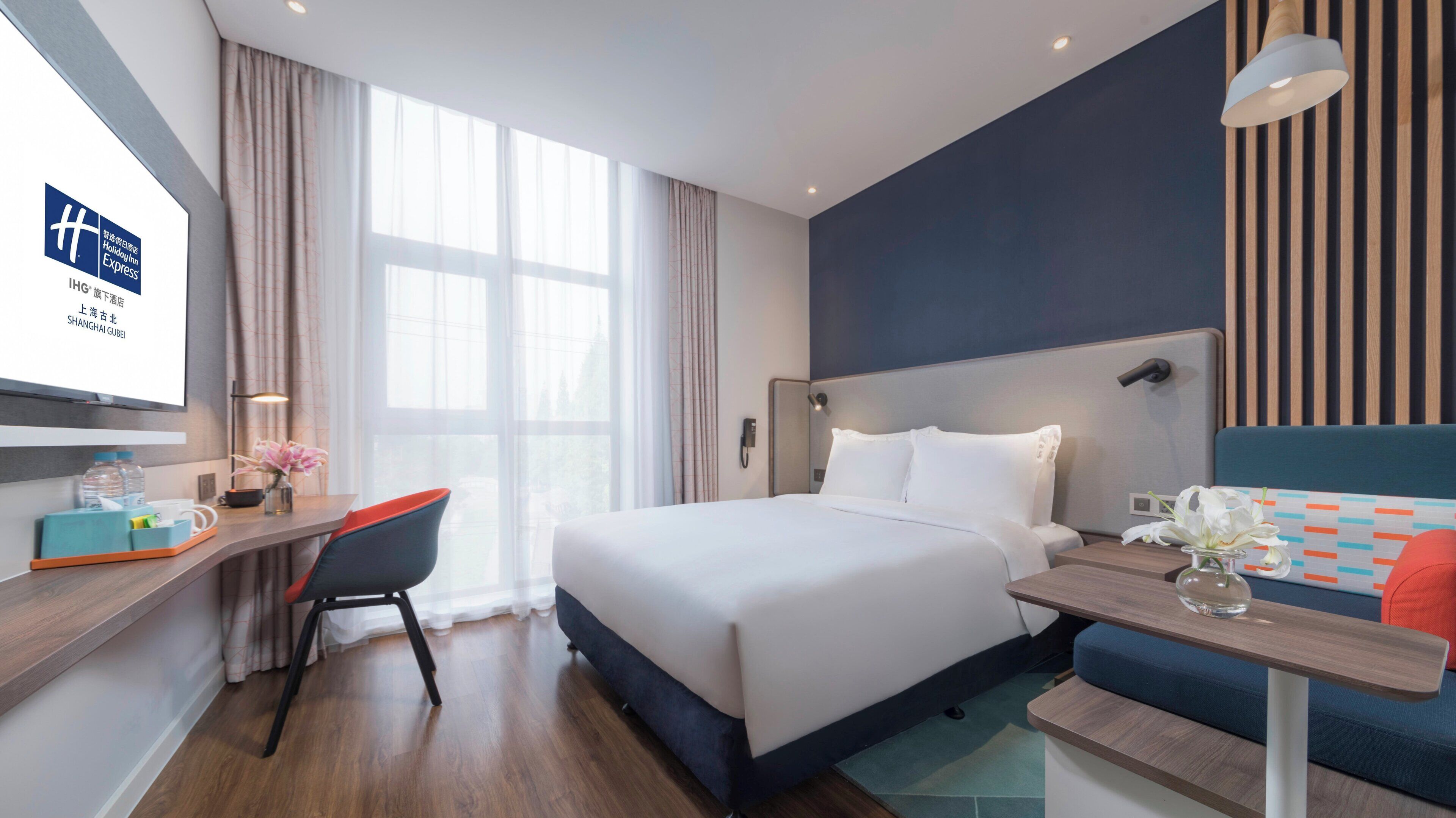 A hotel room with a bed, bedside table, chair, desk, and a large window.