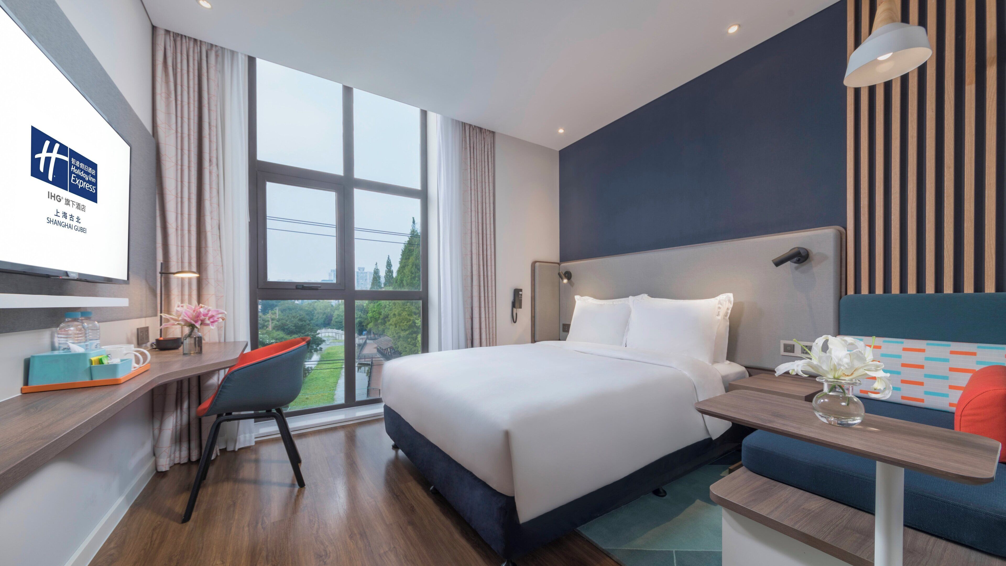 Une chambre d’hôtel avec un grand lit, un bureau avec une chaise, une petite table avec un vase, et une vue sur la verdure à l’extérieur.