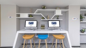 Un espacio de oficina moderno con dos escritorios, cada uno con monitor, teclado y ratón. Hay tres taburetes, uno azul y dos amarillos, y una estantería con objetos decorativos y botellas.
