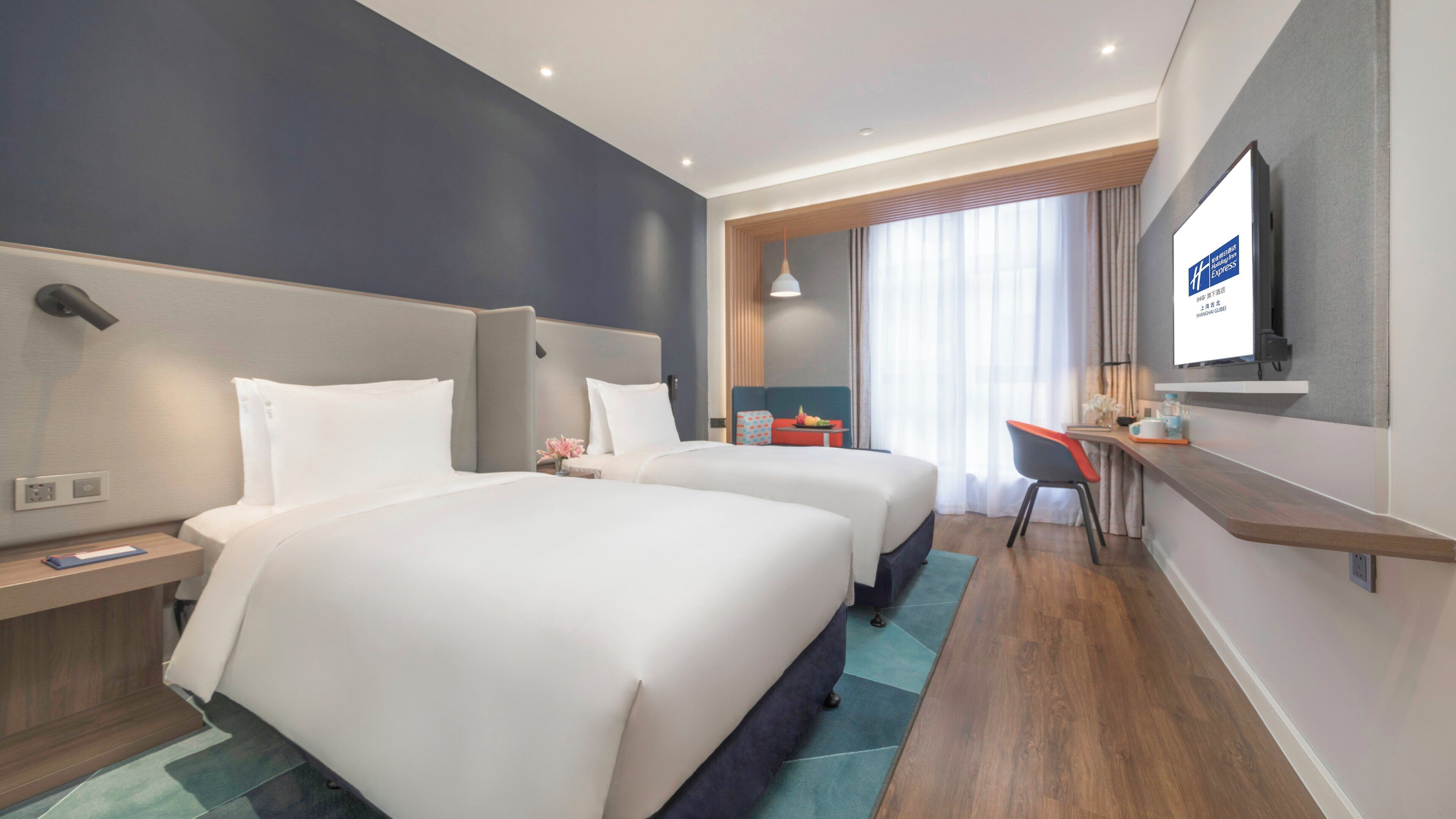 A hotel room with two beds, a desk, a chair, a TV, and a window with curtains.