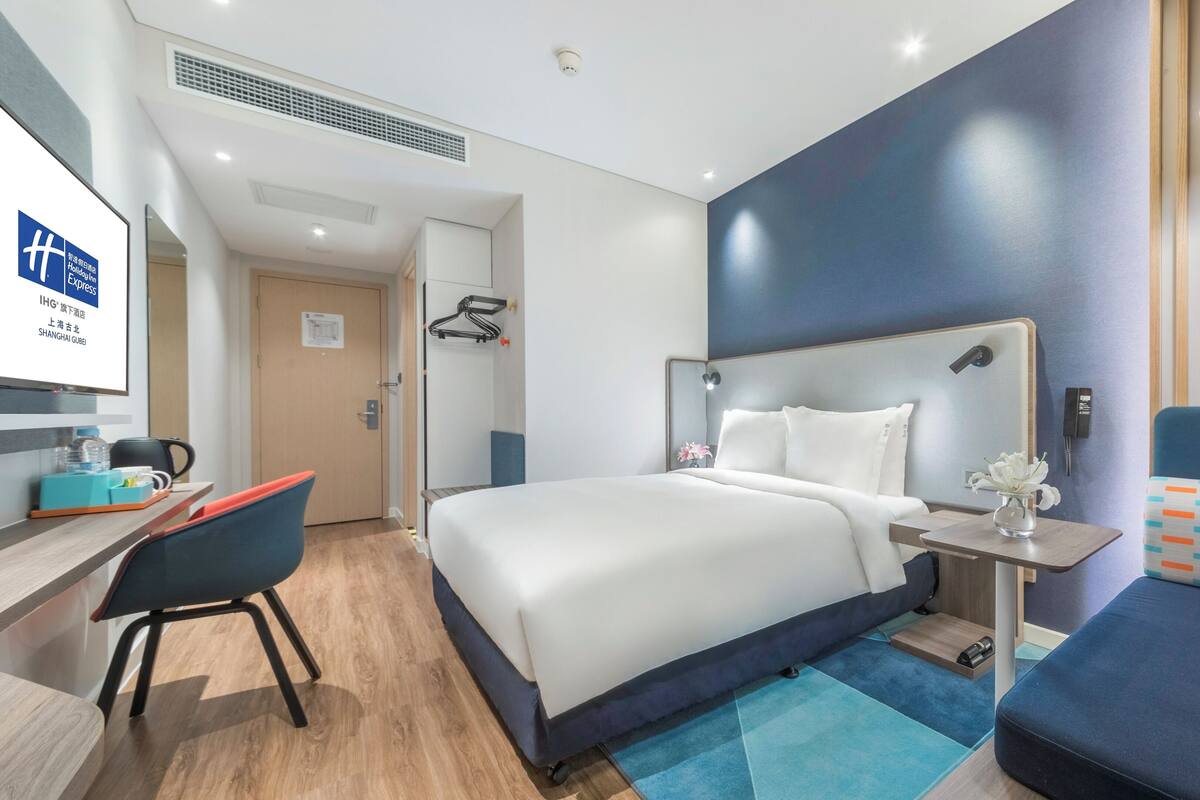 A hotel room with a bed, bedside table, chair, desk, and a small table with a vase.