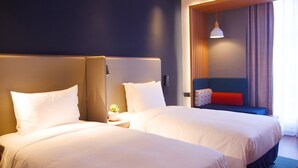 Habitación de hotel con dos camas, una mesita de noche con lámpara, una planta pequeña y una ventana con cortinas.