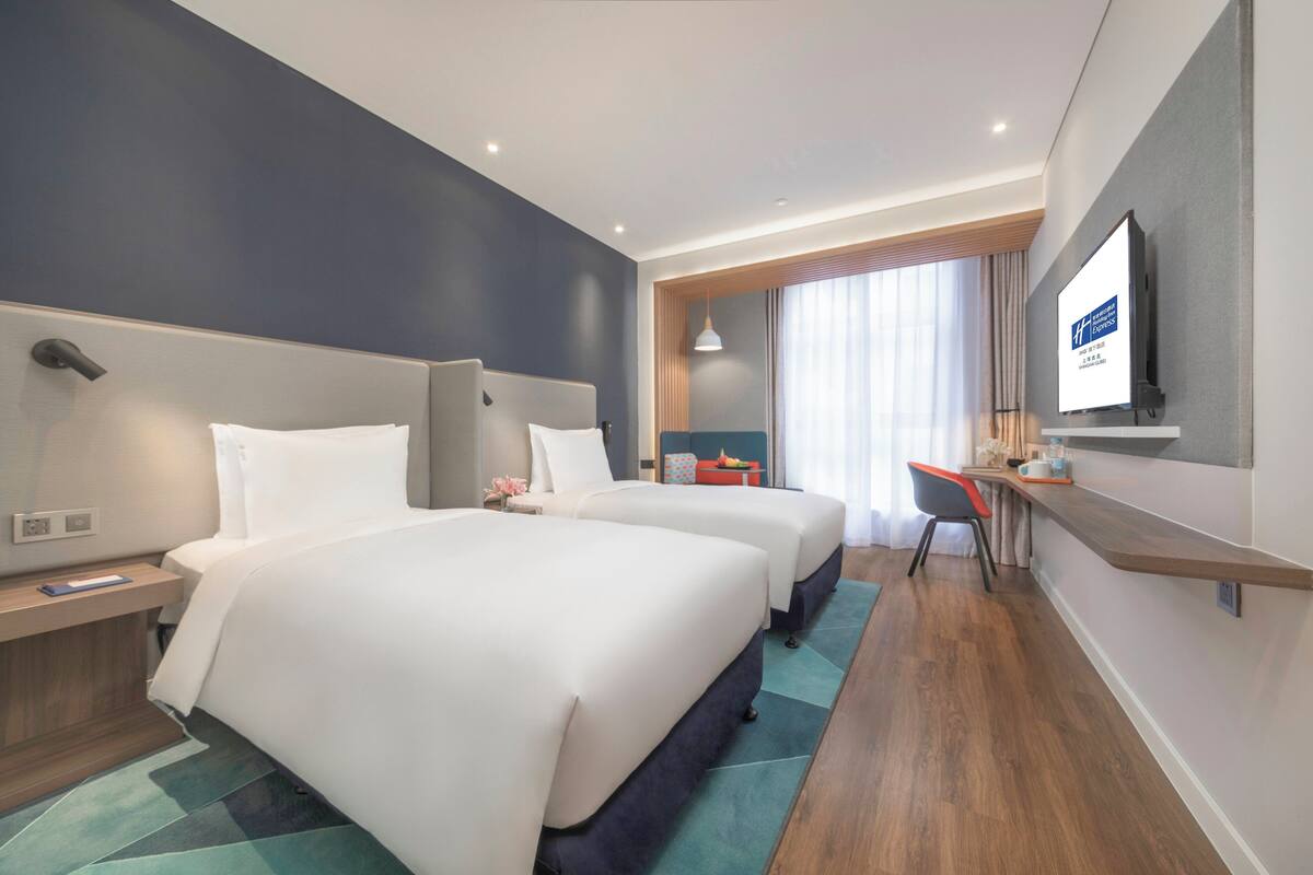 A hotel room with two beds, a desk, a TV, and a window with curtains.