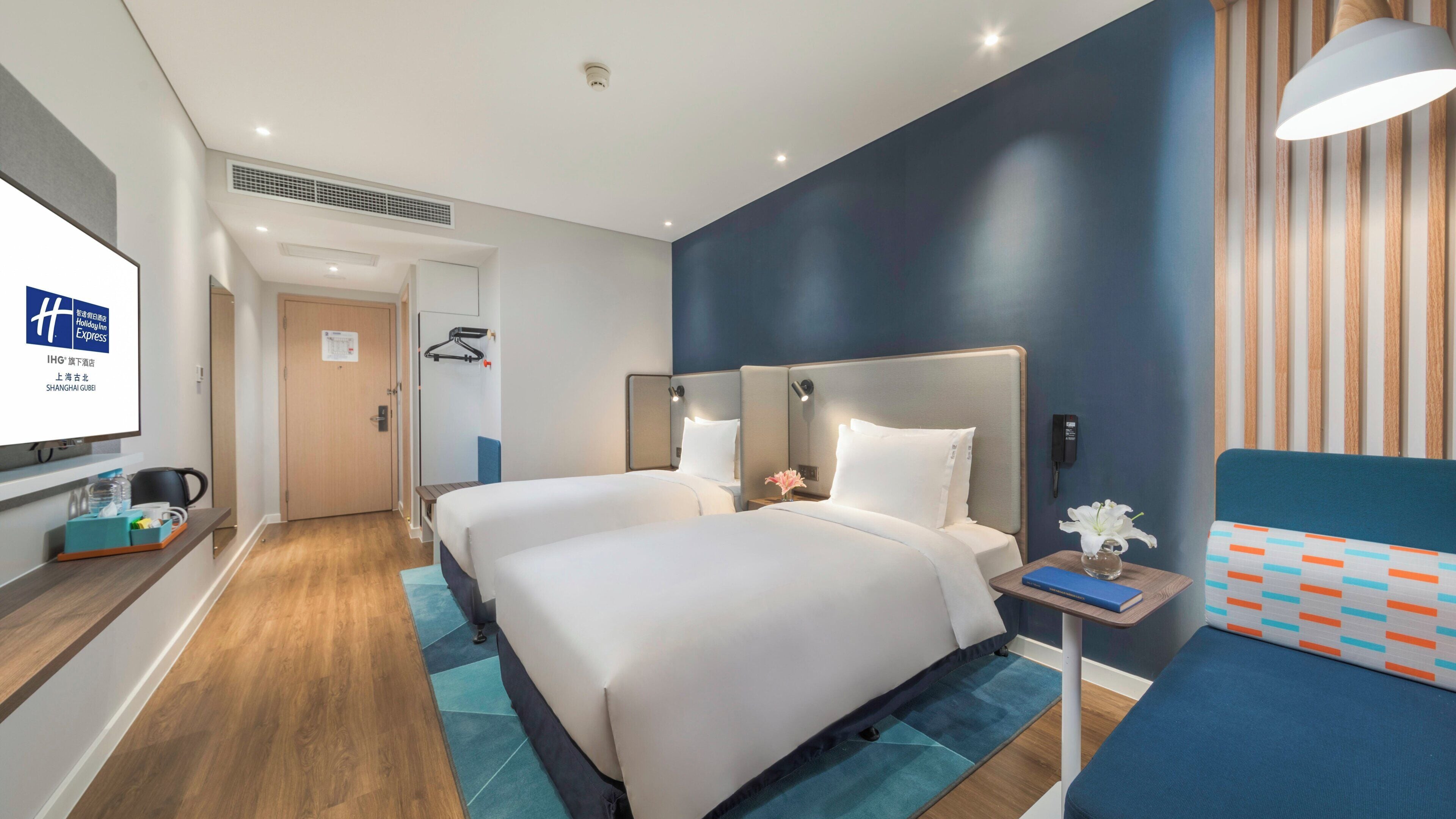 A hotel room with two beds, a desk, a chair, a small table with a vase, and a wall-mounted phone.