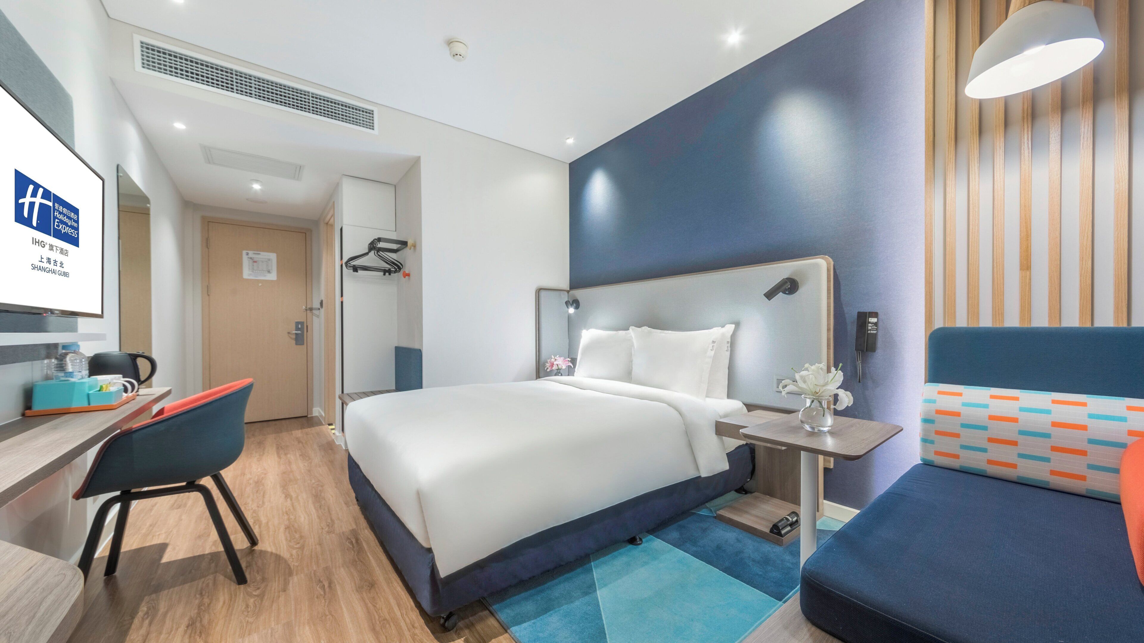 A modern hotel room with a large bed, a desk with a chair, a bedside table with a lamp, and a small table with a vase.