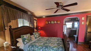 A bedroom with a wooden bed, a ceiling fan, a nightstand, and a view into the kitchen. - Los Gatos Garden Inn Hotel (Los Gatos)