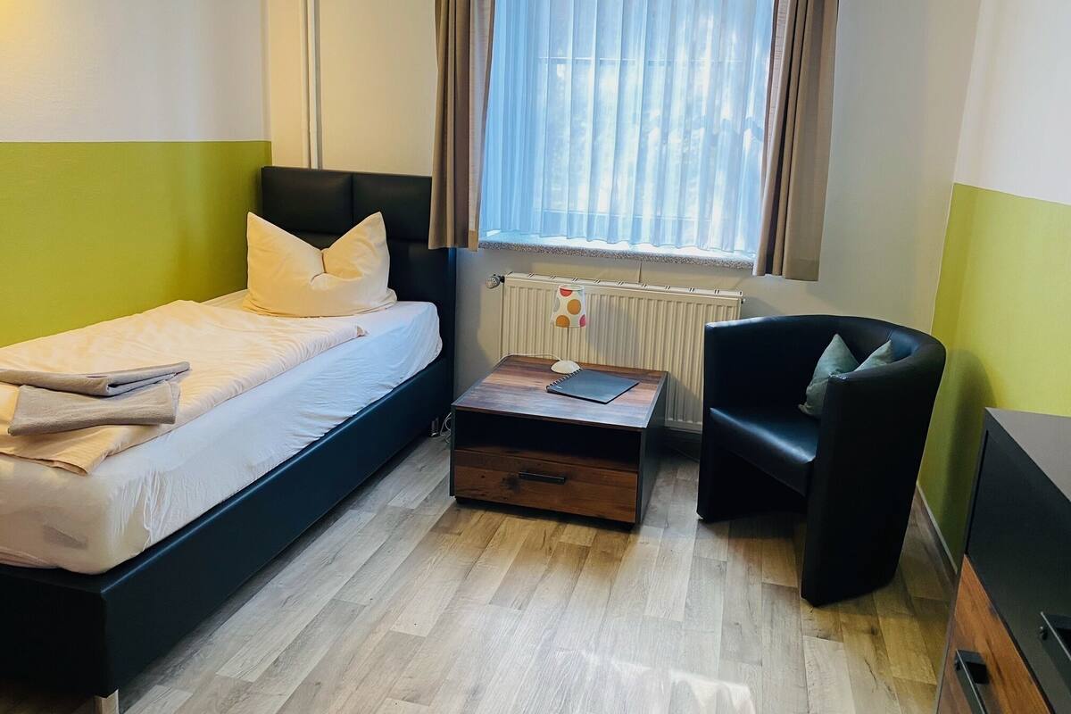 A single bed with a headboard, a bedside table with a lamp, a chair, and a radiator.