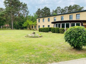 Un bâtiment à deux étages avec une façade en briques, un porche couvert et une pelouse bien entretenue agrémentée d’un élément décoratif en pierre et d’une haie.