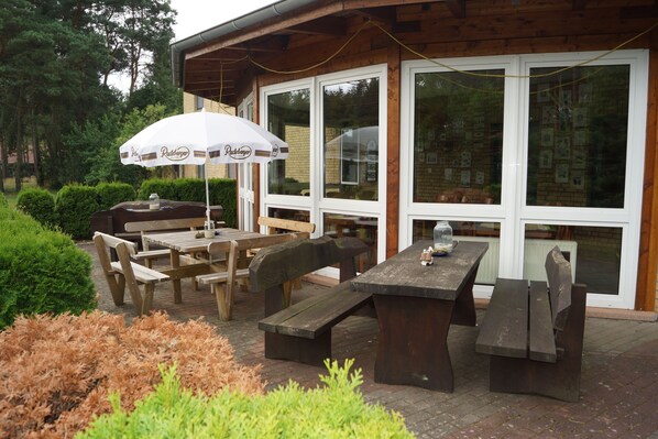 Une terrasse extérieure avec des tables et des bancs en bois, un parasol blanc et un mur en briques.
