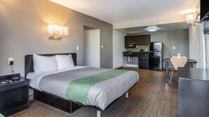 A hotel room with a bed, bedside table, lamp, and a view of the kitchen area. - Studio 6 Little Rock, AR - West (Little Rock)