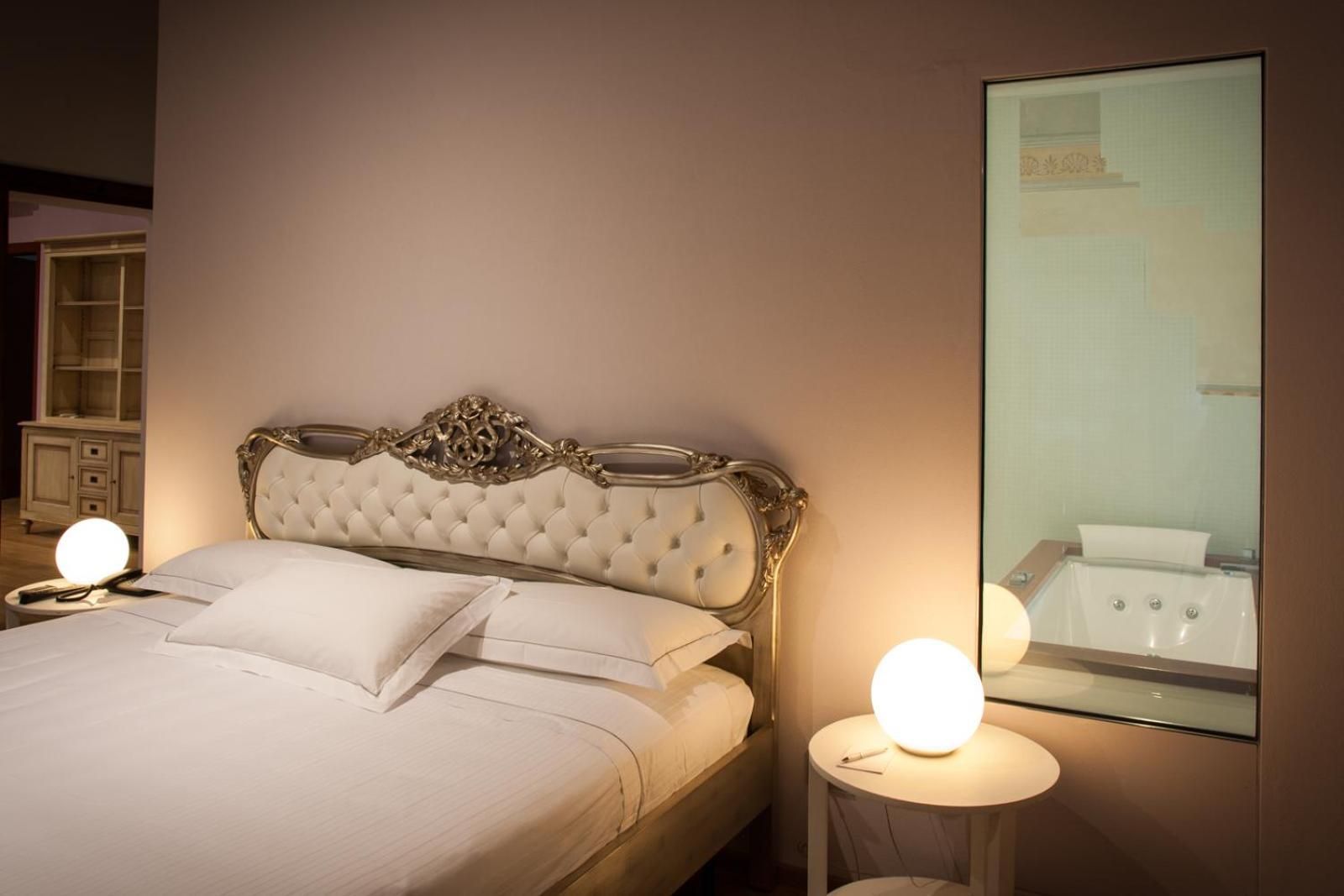 A neatly made bed with a tufted headboard, two bedside lamps, and a large mirror reflecting a bathroom.