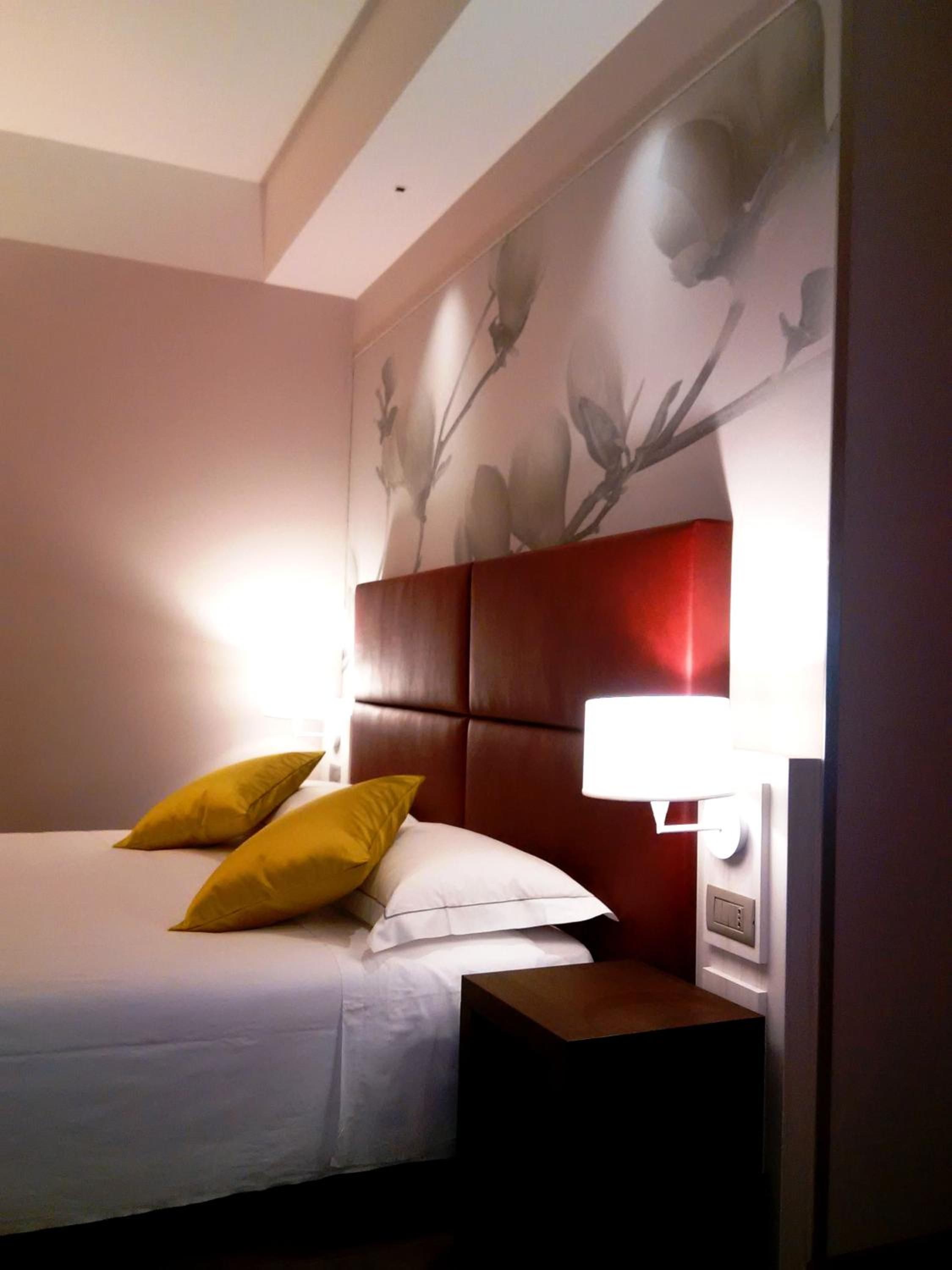 A hotel room with a bed, pillows, a nightstand, a wall-mounted lamp, and a floral wall decoration.