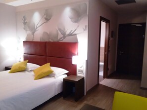 Une chambre d’hôtel avec un lit, des tables de chevet, une commode, une tête de lit rouge et une porte donnant sur une autre pièce.