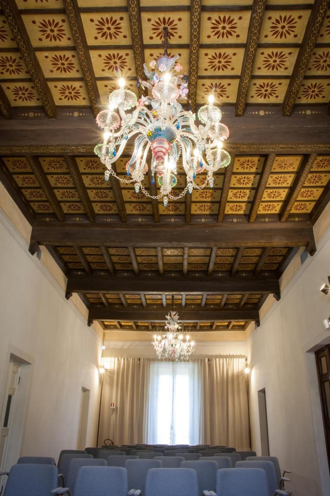 Een kamer met een kroonluchter, een versierd plafond en rijen stoelen.