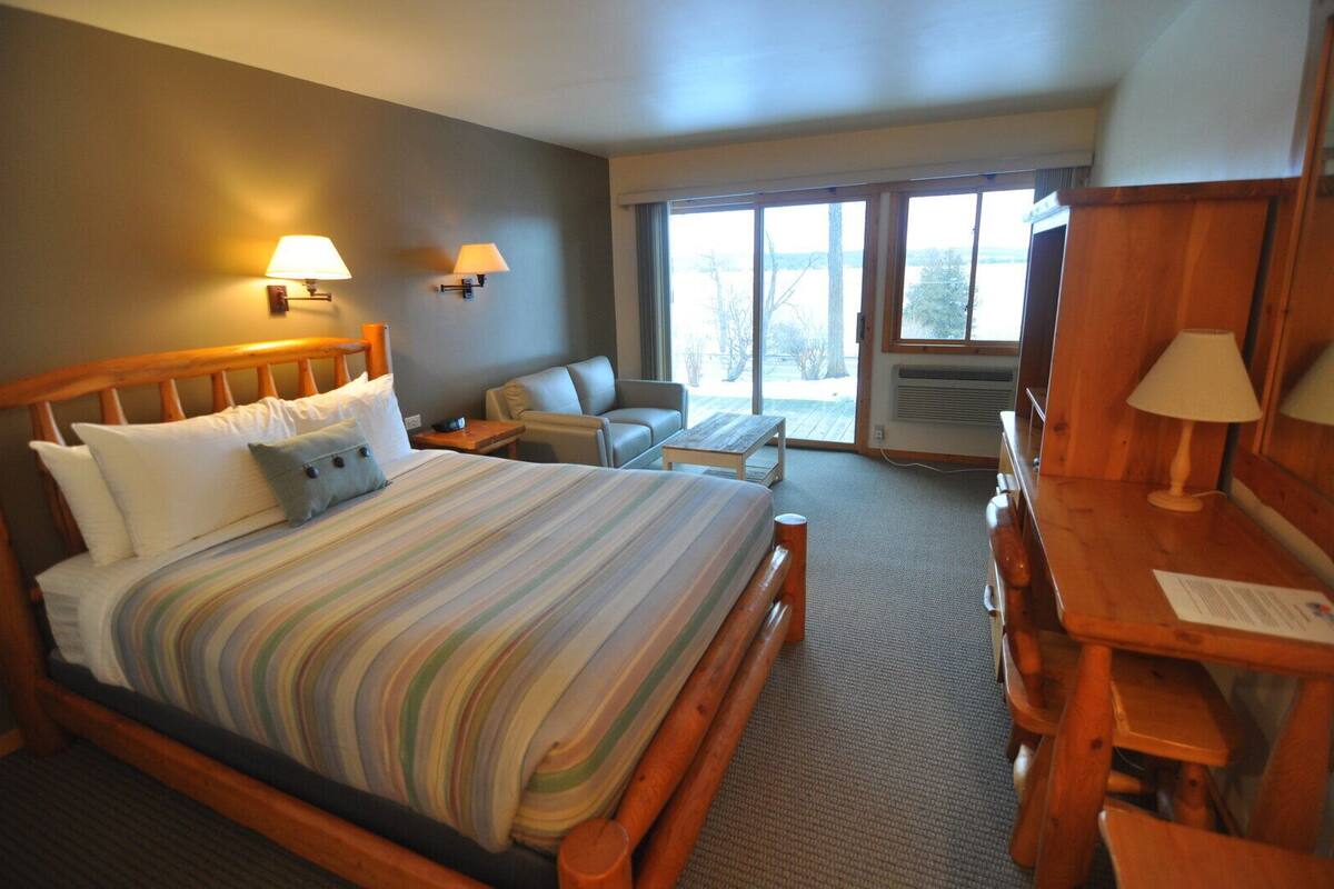 A hotel room with a large bed, a desk, two chairs, a window with a view, and two wall lamps.