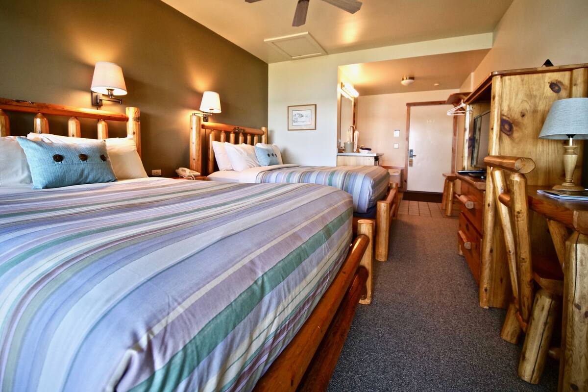 A hotel room with two beds, wooden headboards, a ceiling fan, and a wooden desk with a lamp.