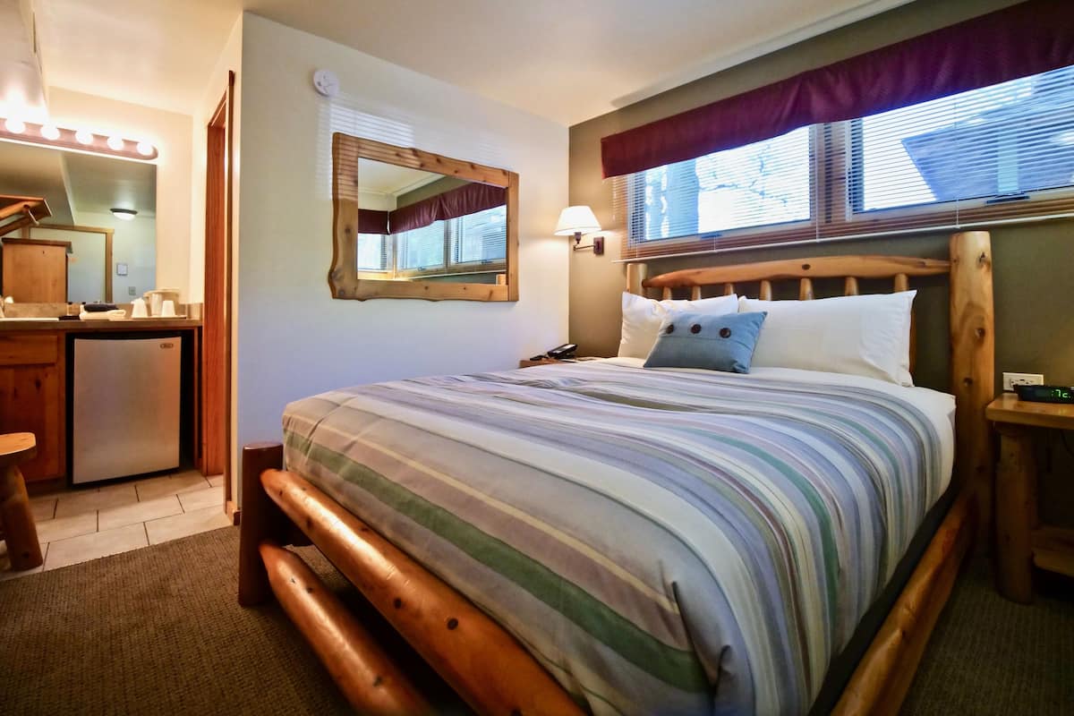 A bedroom with a bed, a wooden headboard, a nightstand, a lamp, a mirror, and a window with blinds.