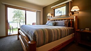 A bedroom with a large bed, a wooden headboard, a nightstand, a lamp, and a view of a lake through a sliding door. - Whaleback Inn (Leland)