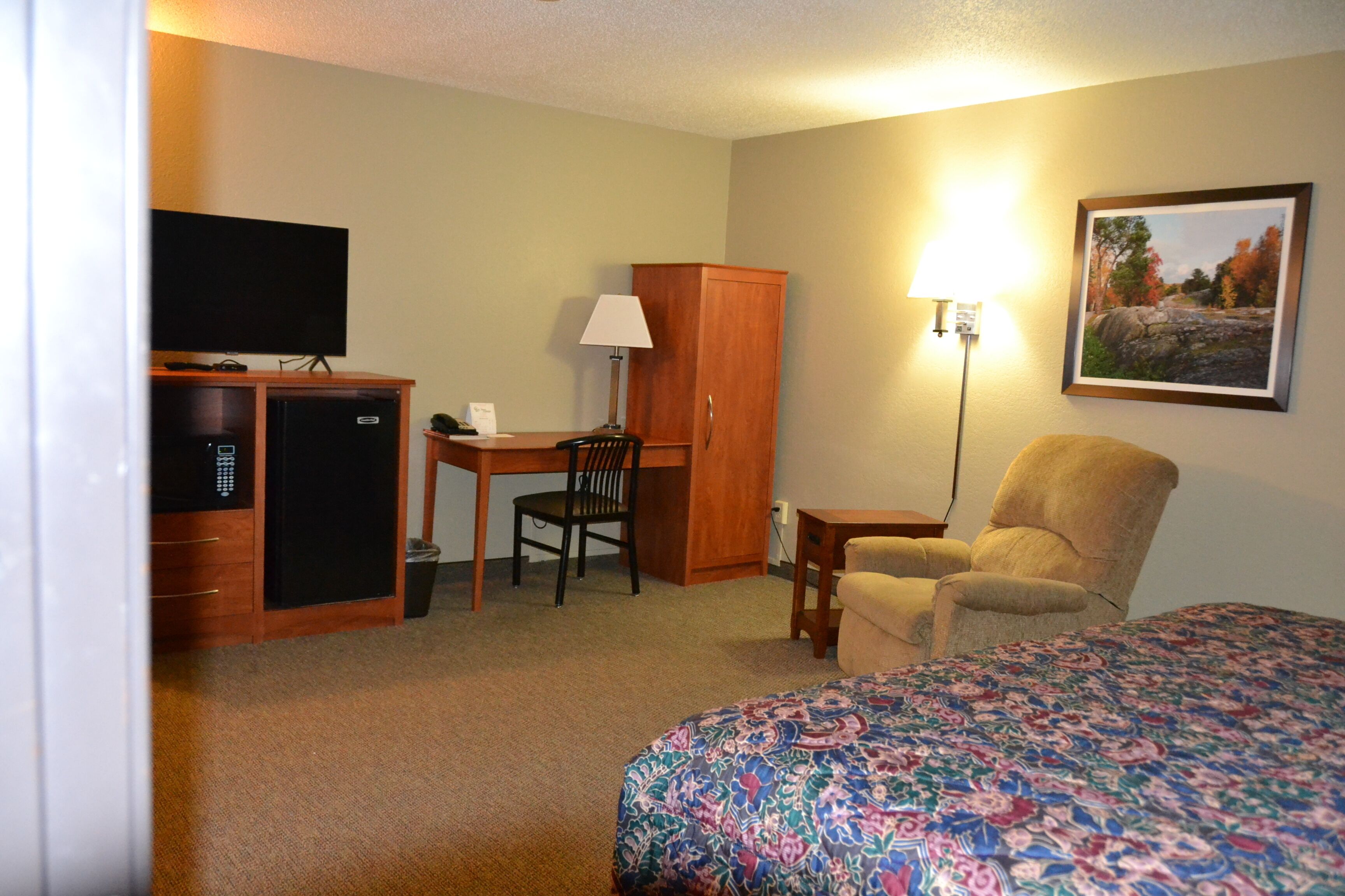 A hotel room with a bed, a desk with a chair, a television, a wardrobe, and a lamp.