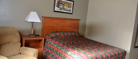 A hotel room with a bed, a chair, a nightstand, a lamp, and a framed picture on the wall.